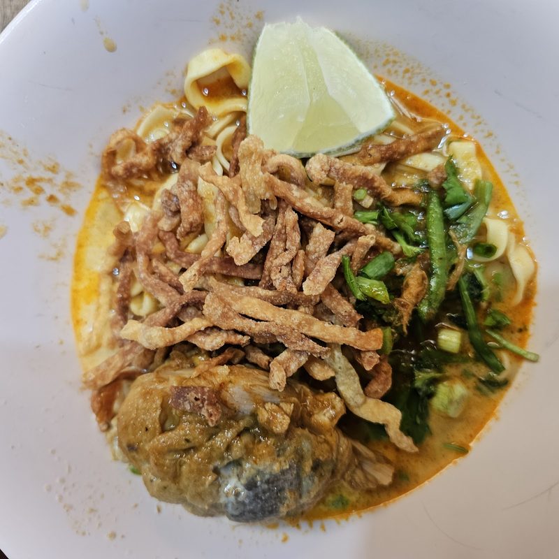 ข้าวซอยไก่มองจากด้านบน เส้นเหลืองในน้ำแกง ไก่ เส้นทอดกรอบ ผัก และมะนาวซีกหนึ่ง