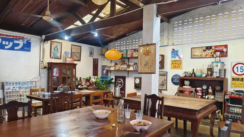 ห้องอาหารในร้าน โต๊ะไม้ พัดลมเพดาน ป้ายและชั้นของเก่า ใต้ผนังบล็อกช่องลม