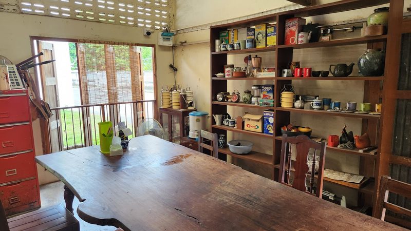 โต๊ะไม้ตัวยาวข้างชั้นวางโถ กาน้ำ และกระป๋องของเก่าในร้าน