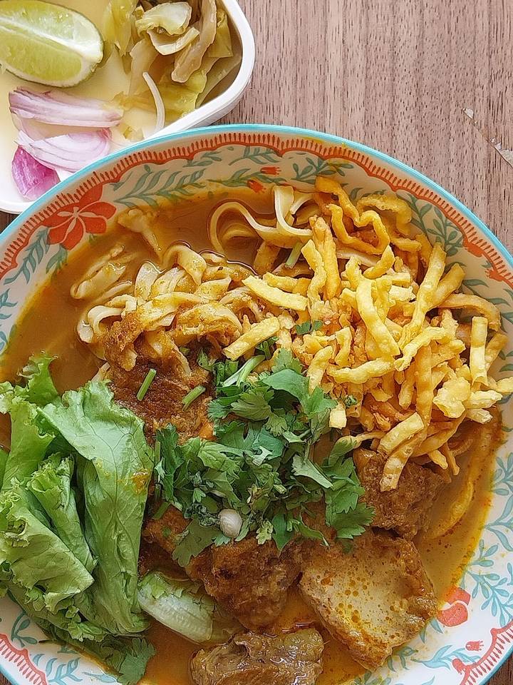 ข้าวซอยในชามลายคราม โรยเส้นกรอบ มีผักกาดหอม ผักชี ผักดอง หอมแดง และมะนาว