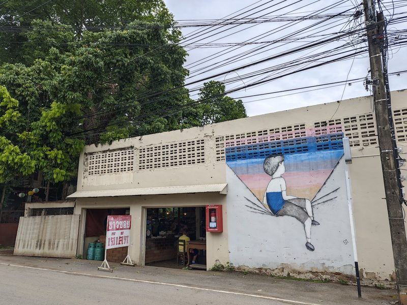 หน้าร้านข้าวซอยเจ๊เล็กบนถนนน้ำคือ ห้องแถวเปิดโล่ง ป้ายตั้งสีแดงขาว และผนังภาพวาดเด็กขนาดใหญ่ข้างร้าน